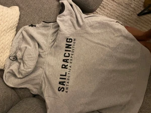 Grå hoodie från Sail Racing - Snygg ljusgrå hoodie från Sail Racing med svart logotyptryck på bröstet och ryggen. Tröjan har hel dragkedja, huva med snörning och ett coolt märke på vänster ärm. Perfekt för chill dagar och har en skön passform.    Storlek 160 barnstorlek ä