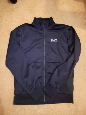 Mörkblå EA7 zipjacka från Armani - Snygg mörkblå zipjacka från EA7 Emporio Armani med vit logga på bröstet och stor tryckt logga på ryggen. Jackan har hög krage, hel dragkedja och ribbade muddar vid ärmar och nederkant. Perfekt för dig som gillar stilren sportig look.