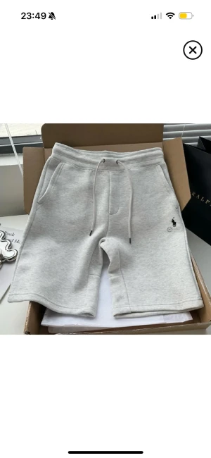 Grå shorts från Ralph Lauren - Snygga grå shorts från Ralph Lauren med klassisk blå logga broderad på framsidan. Modellen har snörning i midjan, fickor på sidorna och en bakficka. Mjukt material som känns chill och passar perfekt till sommaren. Stilren och enkel design. Passar M och L