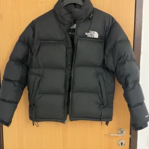 Svart dunjacka från The North Face - Säljer en svart dunjacka från The North Face med broderad logga på bröstet. Jackan har hög krage, dragkedja framtill och justerbara snören i nederkant. Perfekt för kalla dagar och riktigt snygg streetstil.