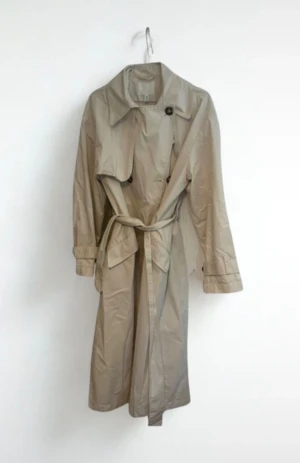 Beige trenchcoat med bälte - Stilren beige trenchcoat med klassisk krage, knappar och bälte i midjan. Jackan har långa ärmar med knäppning vid ärmslut och stora fickor framtill.    Se bilder för skick och eventuella fläckar eller slitningar.  Rökfritt och djurfritt hem!
