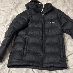 Svart Peak Performance dunjacka - Svart dunjacka från Peak Performance med huva och quiltad design. Jackan har dragkedja framtill, två sidofickor och logga på bröstet. Perfekt för kalla dagar och har en snygg, puffig look som är både praktisk och stilren.