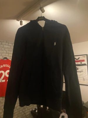 Marinblå zip från Polo Ralph Lauren - Marinblå zip från Polo Ralph Lauren i storlek M. Klassisk design med huva, dragkedja och två fickor framtill. Ikoniska Polo-loggan broderad på bröstet. Perfekt för chill dagar och enkel att matcha med jeans eller joggers.