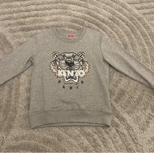 Grå Kenzo sweatshirt med tigertryck - Grå sweatshirt från Kenzo i storlek S med ikoniskt tigertryck och Paris-text framtill. Tröjan har rund hals, ribbade muddar och är långärmad. Perfekt för dig som gillar streetstyle och vill sticka ut med ett snyggt motiv.