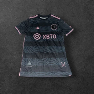 Inter Miami Messi #10 fotbollströja - Säljer en officiell Inter Miami fotbollströja från Adidas med Messi och nummer 10 på ryggen. Tröjan är svart med rosa detaljer och har ett vågigt mönster på framsidan. Kortärmad modell i lätt och ventilerande material, perfekt för match eller träning även som stil. 