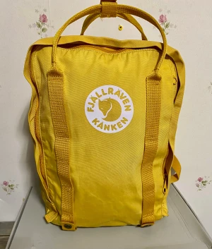 Gul Fjällräven Kånken ryggsäck - Säljer en ikonisk gul Fjällräven Kånken ryggsäck med klassisk rund logga framtill. Väskan har rektangulär form, slitstarka handtag och justerbara axelremmar. Materialet är tålig polyester och väskan har dragkedja upptill för enkel öppning.  Ni kan titta flera bilder, Höjd 38 cm Bredd 27 cm Djup 14 cm