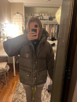 Mörkgrön Fjällräven dunjacka XXXS kan byta - Säljer en brun, riktigt puffig dunjacka från Fjällräven med gul snörning i huvan och stora fickor med guldfärgade knappar. Jackan har broderad Fjällräven-logga på bröstet och dragkedja med knappslå. Perfekt för kalla dagar och har en loose passform. Det är en herr storlek men jag som ofta bär S eller m funkar denna jacka bra på mig! Jag hade även kunnat byta den och betala 2000 ungefär utöver jackan