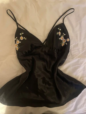 Svart satin linnetopp med broderade blommor - Supersnygg svart linnetopp i satin med tunna axelband och v-ringad front. På bröstet finns broderade vita och gula blommor som ger en extra touch. Toppen är ärmlös och har en mjuk, glansig finish som känns lyxig och trendig.