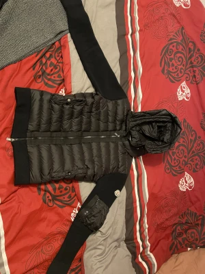 Svart Moncler pufferjacka med huva - Säljer en svart pufferjacka från Moncler med stickade ärmar och huva. Jackan har två fickor med knappstängning, dragkedja framtill och Moncler-logga på ärmen. Materialet är en mix av dun och stickad polyester, perfekt för kyliga dagar.