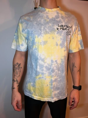 Tie-dye t-shirt från Wasted Paris - Säljer en fet t-shirt från Wasted Paris med tie-dye mönster i ljusblått, gult och vitt. Tryck med logga och tärningar fram, samt stor kors- och textprint på ryggen. Skön passform och tillverkad i mjuk bomull. Perfekt för dig som gillar streetwear och unika prints.