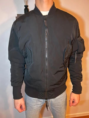 Svart bomberjacka med dragkedja - Svart bomberjacka med klassisk ribbad krage och muddar. Jackan har en rak passform, två snedställda fickor med dragkedja och en extra ficka på ärmen. Materialet är syntet och jackan är helsvart utan mönster. Perfekt för dig som gillar stilren och enkel look. Jackan passar kyligare dagar då den inte är super tunn, dock inte en vinterjacka