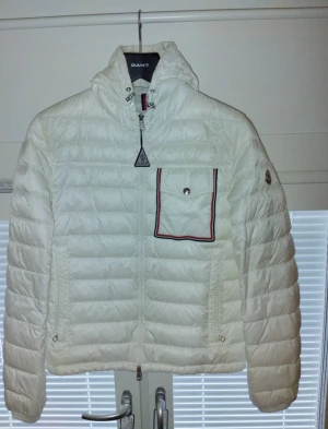 Moncler jacka - Säljer en snygg vit pufferjacka från Moncler med huva och coolt bröstficka med röd och marinblå kant. Jackan har quiltad design, dragkedja framtill och långärmade ärmar. Perfekt för dig som vill ha en stilren och varm jacka med exklusiv känsla. Nypris 14000, inga övriga defekter förutom två små hål där bak. Kom privat för mer frågor!