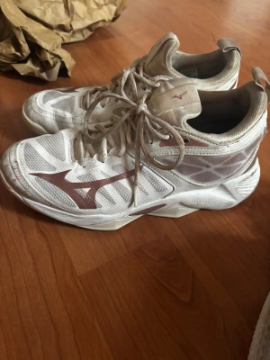 Vita träningskor/innomhusskor från Mizuno  - Säljer ett par vita skor från Mizuno med  ljusrosa detaljer. Skorna har meshpaneler för extra ventilation, snörning och en platt sula. Stora Mizuno-loggan i rosa på sidan ger en sportig vibe. Perfekta för dig som gillar stilrena och bekväma skor. Perfekta inomhus skor för idrotter som ex handboll eller innebandy endast använde en halv säsong, tvättar dem innan jag fraktar dem nypris 1499 