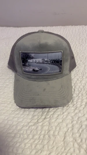 Grå truckerkeps med racingmotiv - Snygg grå truckerkeps från Vernando med mesh på sidorna och baksidan för extra ventilation. Framtill finns ett coolt svartvitt racingmotiv. Justerbar snapback baktill för perfekt passform. Perfekt för dig som gillar motorsport och streetstyle.