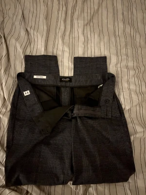 Slim rutig kostymbyxa Jack & Jones - Snygga slim fit kostymbyxor från Jack & Jones i mörkgrått rutigt mönster. Modellen heter Marco och har klassisk midja, bälteshällor och knappstängning. Perfekt för dig som gillar stilrena och moderna detaljer.