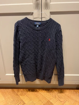 Mörkblå stickad tröja Polo Ralph Lauren - Snygg mörkblå stickad tröja från Polo Ralph Lauren med klassiskt kabelstickat mönster och röd broderad logga på bröstet. Rund halsringning och långa ärmar, perfekt för dig som gillar stilren och tidlös design.