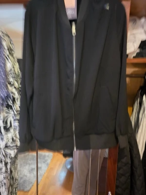 Svart bomberjacka med dragkedja - Svart bomberjacka med klassisk ribbad mudd vid ärmslut och nederkant. Jackan har en rak passform, hel dragkedja framtill och två sidofickor. Materialet är slätt och lätt, perfekt för en chill och stilren look.