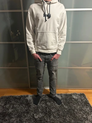 Vit Levi's hoodie med konsttryck - Snygg vit hoodie från Levis med svart logga på bröstet och coolt konsttryck i svart och rosa på ryggen. Klassisk känguruficka framtill, svarta snören i huvan och mjukt bomullsmaterial. Perfekt för dig som gillar streetwear och unik design.