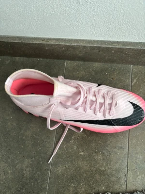 Nike mercurial 40,5 rosa fotbollsskor - Säljer ett par snygga fotbollsskor från Nike i ljusrosa med svarta detaljer och en stor swoosh på sidan. Skorna har snörning och en platt sula med rosa dobbar för gräsplan. Ovandelen är i syntetmaterial och insidan har en rosa ton. Perfekt för dig som vill sticka ut på planen.