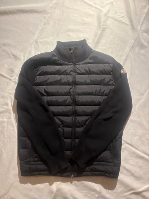 Svart cardigan från Moncler - Säljer en svart cardigan från Moncler med quiltad framsida och stickade ärmar samt rygg. Jackan har hög krage, dragkedja framtill och Moncler-logga på ärmen. Perfekt för dig som gillar stilren design och exklusiva märken. 