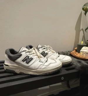 New Balance 550 vita sneakers - Säljer ett par New Balance 550 sneakers i vitt med grå och svarta detaljer. Klassisk chunky siluett med rund tå, snörning och platt sula. Ovandelen är i skinn med perforerade paneler och tydlig New Balance-logga på sidan. Perfekt för dig som gillar retrostil och streetwear.