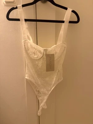 Vit spetsbody - Säljer en vit body med breda axelband och vacker spets över hela plagget. Modellen har markerade kupor och en feminin siluett. Perfekt för dig som vill ha en chic och trendig topp med lite edge. 