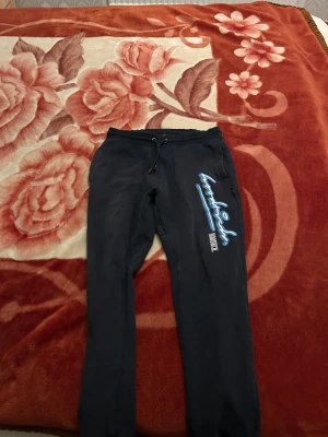 Svarta mjukisbyxor från Hoodrich  - Svarta mjukisbyxor från Hoodrich i storlek S med blått och vitt tryck längs benet. Byxorna har snörning i midjan och ribbade muddar vid bensluten. Mjukt material som är perfekt för chill eller träning. Snygg streetstil med coola detaljer.