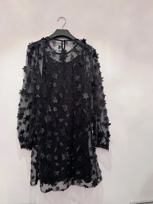 Klänning  - Supersnygg svart klänning i mesh med 3D-blommor över hela och diskreta glittriga prickar. Klänningen har långärmade transparenta ärmar och rund hals med knapp i nacken. Perfekt för dig som vill sticka ut med coola detaljer och unik look. Perfekt till nyår. 