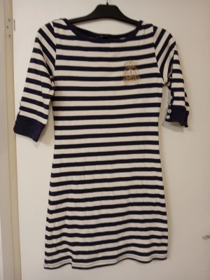 Randig klänning från Henri Lloyd - Snygg kortklänning från Henri Lloyd med marinblå och vita ränder. Klänningen har trekvartsärm med knappar vid ärmslutet och ett broderat märke på bröstet. Perfekt för en avslappnad men stilren look.