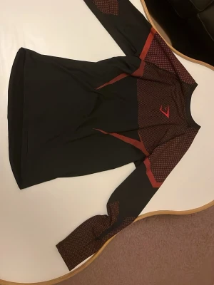 Svart långärmad träningströja Gymshark - Snygg svart långärmad träningströja från Gymshark med röda detaljer och mönster på axlar och ärmar. Tillverkad i ett stretchigt funktionsmaterial som andas, perfekt för gymmet eller löprundan. Tight passform och sportig look.