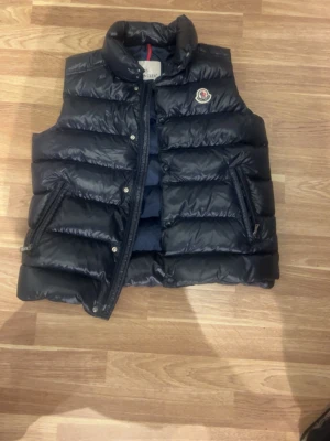 Mörk blå dunväst från Moncler - Snygg svart dunväst från Moncler med hög krage och två dragkedjefickor framtill. Västen har ett klassiskt Moncler-märke på bröstet och quiltad design. Insidan har en unik serietidningsetikett och märkning. Perfekt för lager-på-lager och streetstyle.