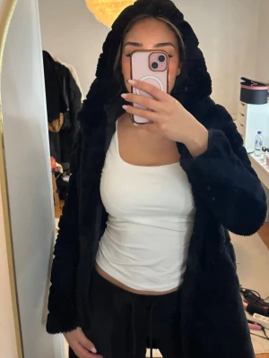 Svart faux pälsjacka med huva - Snygg svart pälsjacka med huva och lång ärm. Jackan har en rak passform och är gjord i mjuk fuskpäls som känns lyxig och varm. Perfekt för att hålla sig varm under kyliga dagar och ger en trendig look.