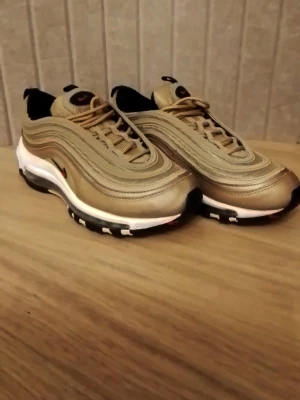 Nike Air Max 97 Gold sneakers - Säljer ett par Nike Air Max 97 i metallic guld med svarta och vita detaljer. Skorna har den ikoniska vågiga designen, synlig Air-sula och röd Swoosh-logga på sidan. Ovandelen är i syntet och mesh för en sportig look. Perfekta för dig som vill sticka ut.