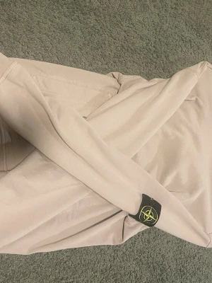 Beige hoodie från Stone Island - Snygg beige hoodie från Stone Island med den klassiska svarta logotypen med gul och grön kompass på ärmen. Hoodien har en avslappnad passform och är tillverkad i mjukt bomullsmaterial. Perfekt för en stilren och trendig look.