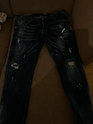 Mörkblå distressed jeans med dragkedja - Säljer ett par mörkblå jeans från Dsquared2 med slitningar, färgstänk och en cool dragkedjedetalj vid fickan. Jeansen har en rak passform och klassisk femficksdesign. Perfekta för dig som gillar en edgy streetstil.