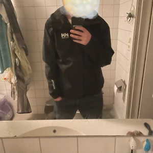 Svart regnjacka från Helly Hansen - Svart regnjacka från Helly Hansen med dragkedja framtill och hög krage. Jackan har en enkel, sportig design och är tillverkad i ett lätt syntetmaterial som står emot regn. Perfekt för blöta dagar och enkel att matcha med olika outfits.