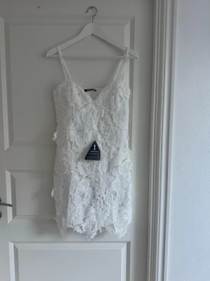 Vit spetsklänning med blommönster - Supersöt vit kort klänning med tunna axelband och vackra spetsdetaljer i blommönster. Klänningen har en figurnära passform och är perfekt för sommarens alla tillfällen. Materialet känns lätt och luftigt.