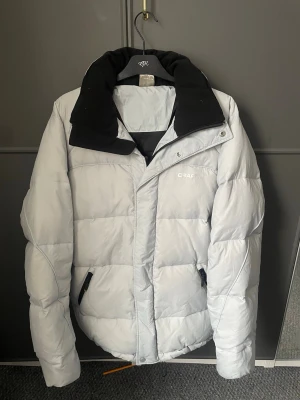 Ljusgrå dunjacka från Craft, M - Snygg ljusgrå dunjacka från Craft med svart krage och dragkedja. Jackan har quiltad design, två fickor med dragkedja och hög krage för extra värme. Perfekt för kalla dagar och enkel att matcha med olika outfits.