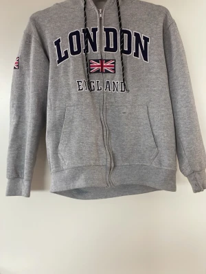 Grå hoodie med London-tryck - Snygg grå hoodie med dragkedja och huva, prydd med texten 'LONDON ENGLAND' och en Union Jack-flagga på bröstet. Svarta snören med vita detaljer och fickor framtill. Perfekt för dig som gillar streetwear och vill ha en chill look.