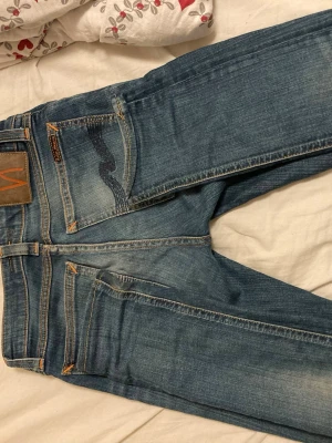 Blå jeans från Nudie Jeans - Snygga blå jeans från Nudie Jeans med klassisk femficksdesign och kontrastsömmar. Jeansen har en normal passform och är tillverkade i slitstarkt bomullsdenim. Perfekta för dig som gillar en tidlös och avslappnad stil.