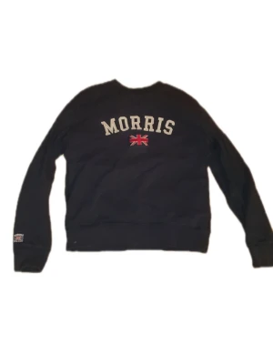  sweatshirt från Morris - Svart sweatshirt från Morris med stor logga och brittisk flagga broderad på bröstet. Klassisk rund halsringning och ribbade muddar vid ärmslut och nederkant. Perfekt för en avslappnad och stilren look.