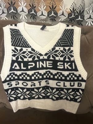 Vit stickad väst med mönster - Säljer en vit stickad väst med svart vintermönster och texten 'ALPINE SKI SPORTS CLUB' framtill. Västen har v-ringning och breda ribbade kanter. Perfekt för dig som gillar retro och sportig stil.