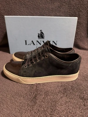 Blå/gröna lanvins med dustbag  - Stilrena svarta sneakers från Lanvin med ovandel i mocka och detaljer i skinn. Skorna har snörning och en beige platt sula som ger en clean look. Perfekta för dig som gillar klassisk design med lyxig känsla.