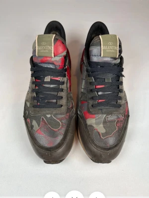 Valentino Rockrunner camo sneakers - Snygga Valentino Rockrunner sneakers med camouflage-mönster i rött, svart och grått. Skorna har svarta snören, rund tå och detaljer i mocka och skinn. Ikoniska nitar baktill och logga på plösen. Perfekta för dig som vill sticka ut.