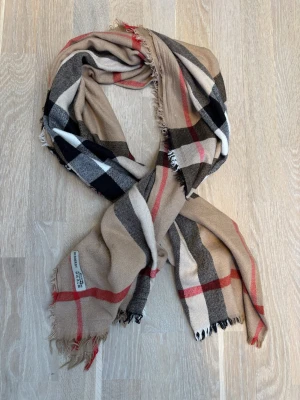Rutig halsduk från Burberry i ull & kashmir - Klassisk rutig halsduk från Burberry i beige, svart, röd och vit. Tillverkad i mjuk ull och kashmirblandning. Tidlös design med fransade kanter och ikoniskt rutmönster. Perfekt accessoar för att lyfta vilken outfit som helst.