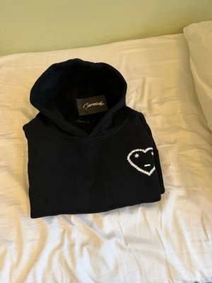 Carsicko hoodie - Säljer en svart hoodie från Carsicko med ett vitt broderat hjärta och ansikte på bröstet. Hoodien har huva, ribbade muddar och en klassisk passform. Perfekt för dig som gillar streetwear och vill ha något stilrent men ändå unikt.