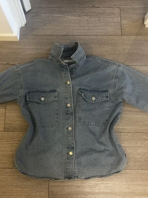 Blå jeansjacka - Blå oversized jeansjacka från Ellos med två stora bröstfickor och silverfärgade knappar framtill. Jackan har en rak passform och lång ärm, inga synliga defekter, storlek 36/40