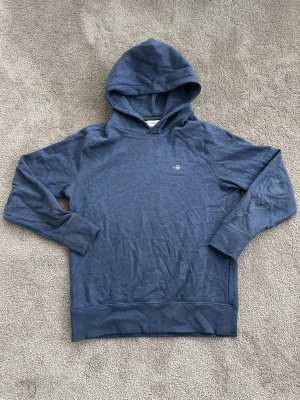 Blå hoodie från Gant - Snygg blå hoodie från Gabr med minimalistisk logga på bröstet. Hoodien har klassisk passform, långa ärmar och ribbade muddar vid ärmslut och nederkant. Vid frågor är det bara att slänga iväg ett meddelande!