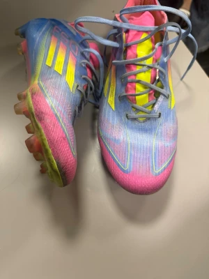 Adidas F50 färgglada fotbollsskor - Säljer ett par Adidas F50 fotbollsskor i en riktigt färgstark mix av blått, rosa och gult. Skorna har snörning, låg profil och är tillverkade i syntetmaterial för lätt känsla på planen. Perfekta för dig som vill sticka ut på fotbollsplanen.