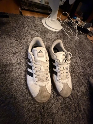 Adidas Samba vita sneakers - Klassiska Adidas Samba sneakers i vitt med svarta ränder och grå detaljer. Skorna har snörning, rund tå och en brun gummisula. Tillverkade i syntet och mocka för en retro och sportig look. Perfekta för dig som gillar tidlös streetstyle.